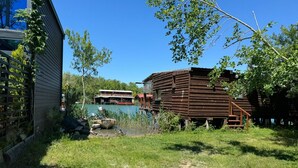 Una cabaña rústica con un muelle que se adentra en un lago, rodeada de exuberante vegetación y un cielo azul despejado.