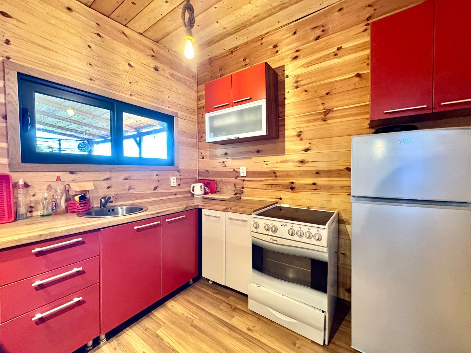 A compact kitchen with wooden walls, a window, a microwave, a stove, and a refrigerator.