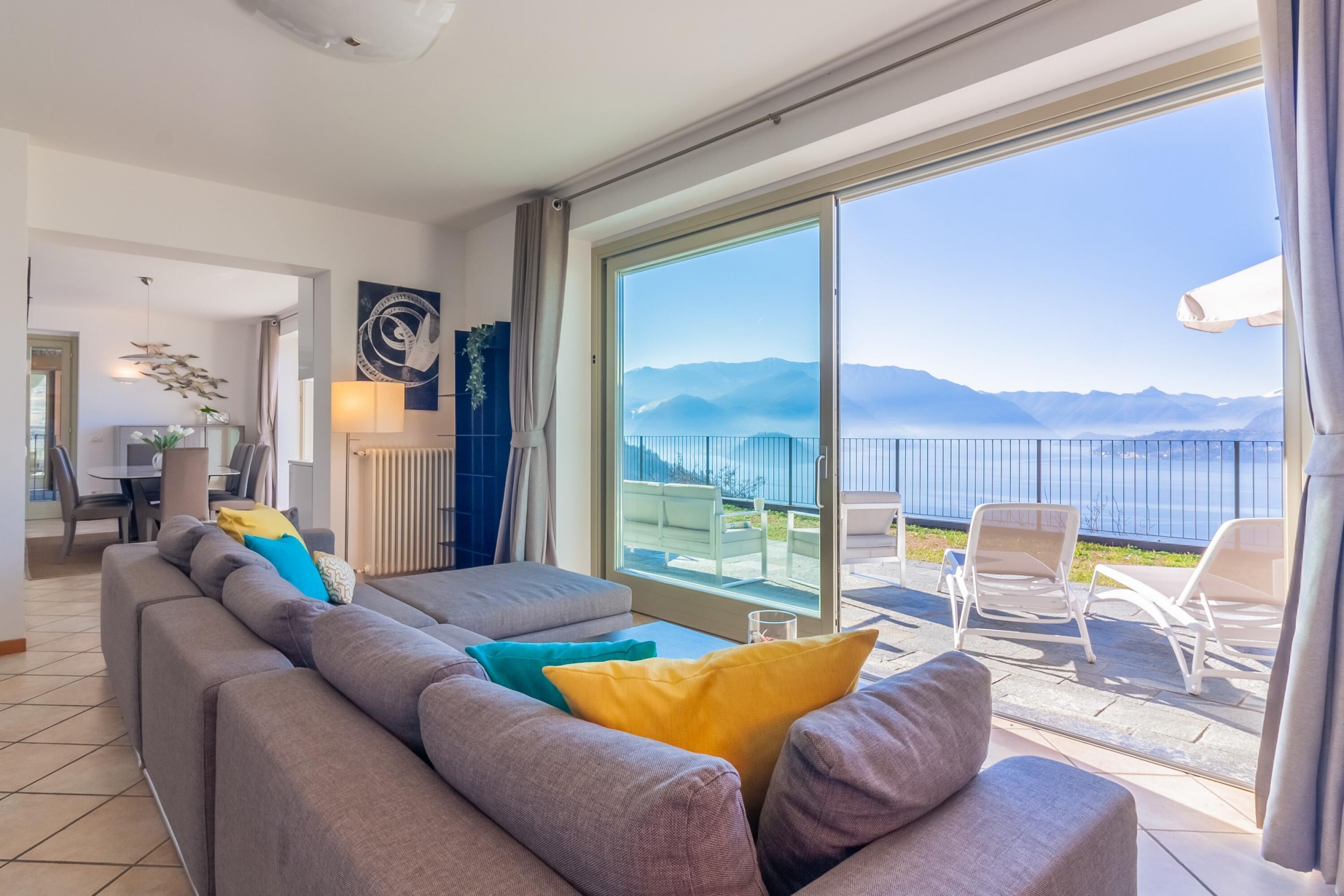 A modern living room with a grey sofa, colorful cushions, and a large sliding glass door leading to a balcony with a view of mountains and a body of water.