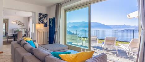 A modern living room with a grey sofa, colorful cushions, and a large sliding glass door leading to a balcony with a view of mountains and a body of water.