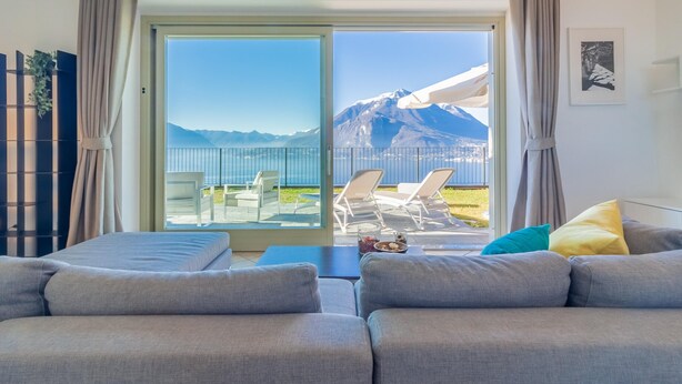 A modern living room with a sofa and a dining area. A sliding glass door leads to an outdoor patio with a view of mountains. - The Patio Over Varenna (Perledo)