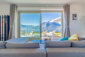 A modern living room with a sofa and a dining area. A sliding glass door leads to an outdoor patio with a view of mountains. - The Patio Over Varenna (Perledo)