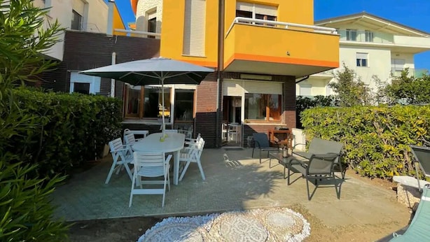 A modern two-story house with a curved facade, outdoor seating area with white chairs and a table, and a small patio with a table and chairs.