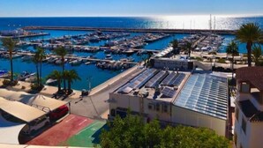 Un puerto deportivo con barcos amarrados, una carretera que lleva a un edificio con paneles solares, y un cielo azul despejado.