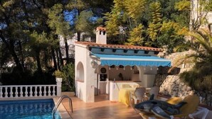 Zona de piscina con solárium, mobiliario exterior y un pequeño edificio con tejado de baldosas.
