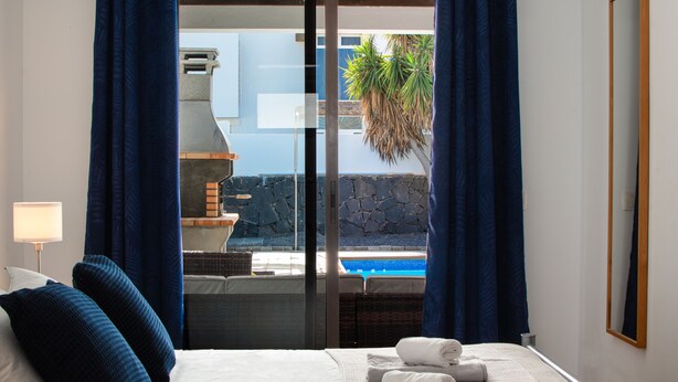 A hotel room with a bed, pillows, a nightstand, a lamp, a view of an outdoor pool, and a stone wall. - Casa Marlay Spacious Villa Pool, BBQ & Fast Wi-fi Sun all day (Playa Blanca)