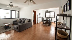 A spacious living room with a grey sofa, a ceiling fan, and a dining area with a table and chairs. - Casa Marlay Spacious Villa Pool, BBQ & Fast Wi-fi Sun all day (Playa Blanca)