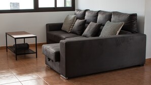 A modern living room with a dark grey sofa, multiple cushions, a small side table, and a window with blinds. - Casa Marlay Spacious Villa Pool, BBQ & Fast Wi-fi Sun all day (Playa Blanca)