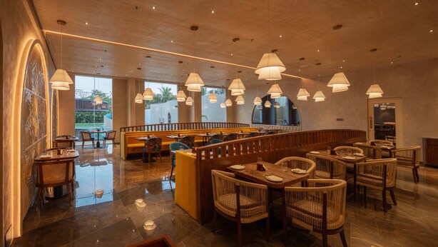 A spacious restaurant with high ceilings, multiple dining areas, and a central seating area with a view of the outdoors. - Palm Springs Resort (Nashik)