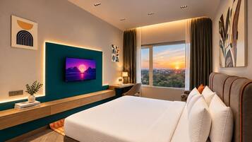 A modern hotel room with a large bed, a TV mounted on the wall, a desk with a lamp, and a view of the city at sunset.
