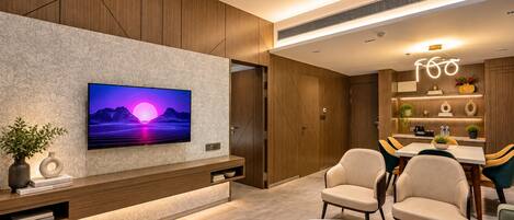 Una sala moderna con televisor de pantalla plana montado en una pared con textura, una zona de comedor con mesa y sillas, y una cocina integrada con estantes que exhiben objetos decorativos.