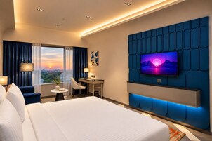 Habitación de hotel con una cama grande, un escritorio, una silla, un televisor montado en una pared azul y vistas a la ciudad al atardecer.