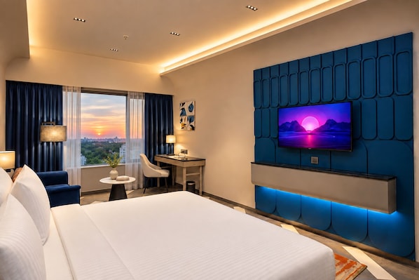 Habitación de hotel con una cama grande, un escritorio, una silla, un televisor montado en una pared azul y vistas a la ciudad al atardecer.