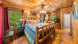 A cozy cabin room with a wooden ceiling, a bed with a maple leaf pillow, a nightstand, a window with blinds, and a door. - Fallin' in Love: Lose Your Heart in the Smokies (Sevierville)