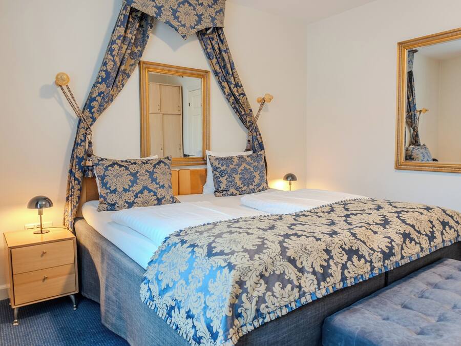 A bedroom with a bed, a nightstand, a bench, a mirror, and curtains.