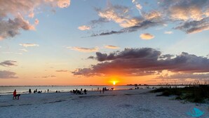 Una spiaggia con persone che camminano e siedono, un molo sullo sfondo e un tramonto.
