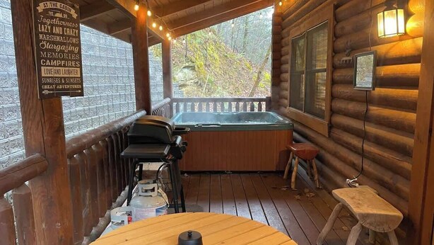 A wooden deck with a hot tub, a table, and a sign listing amenities like 'marshmallows' and 'campfires'. - Cabin for 4 • Hot Tub • Jetted Tub • Arcade Loft (Sevierville)