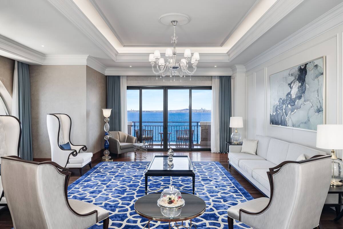 A spacious living room with a large window, a chandelier, and a view of the sea.