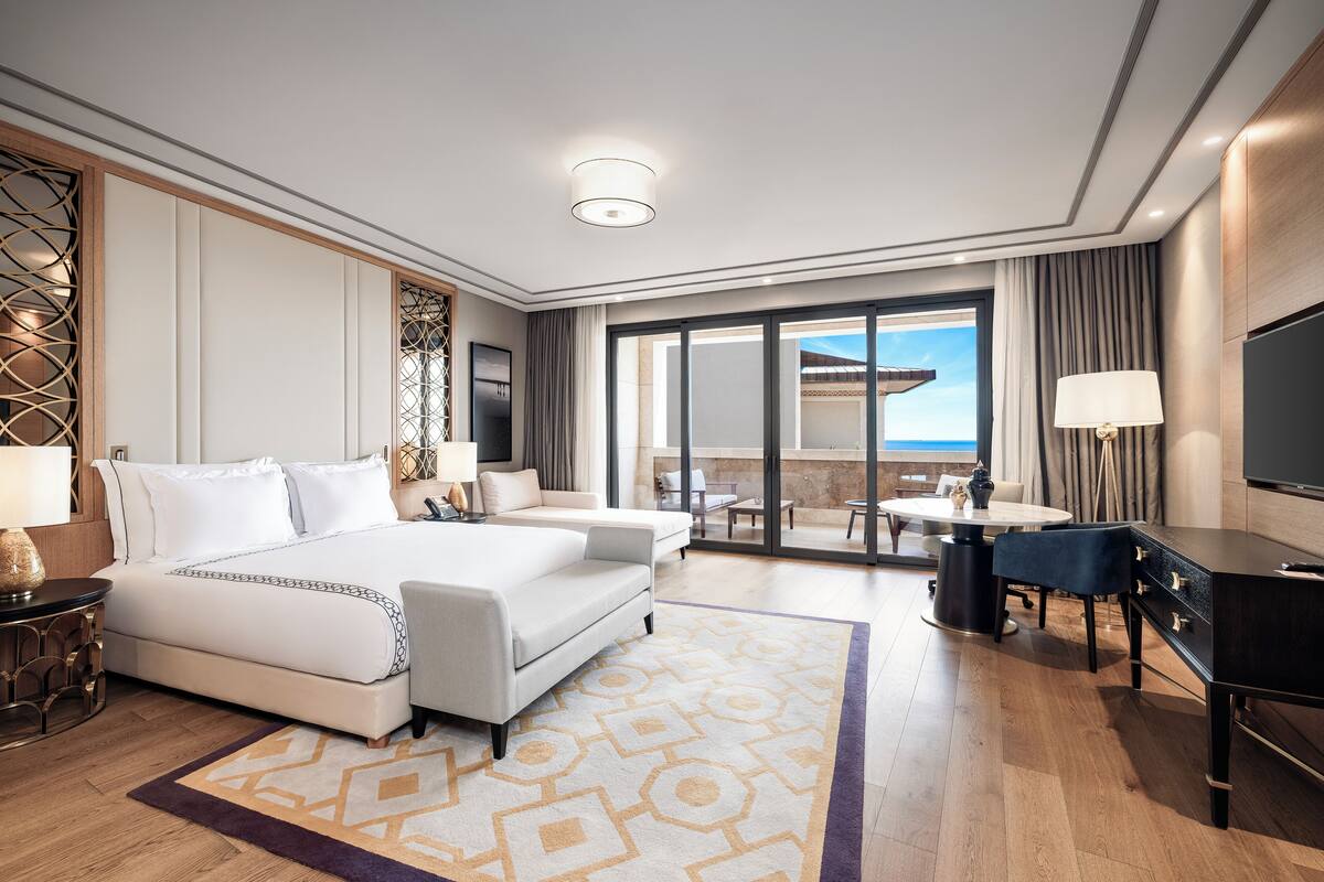 A modern hotel room with a large bed, a desk, a chair, a TV, and a balcony with a view.