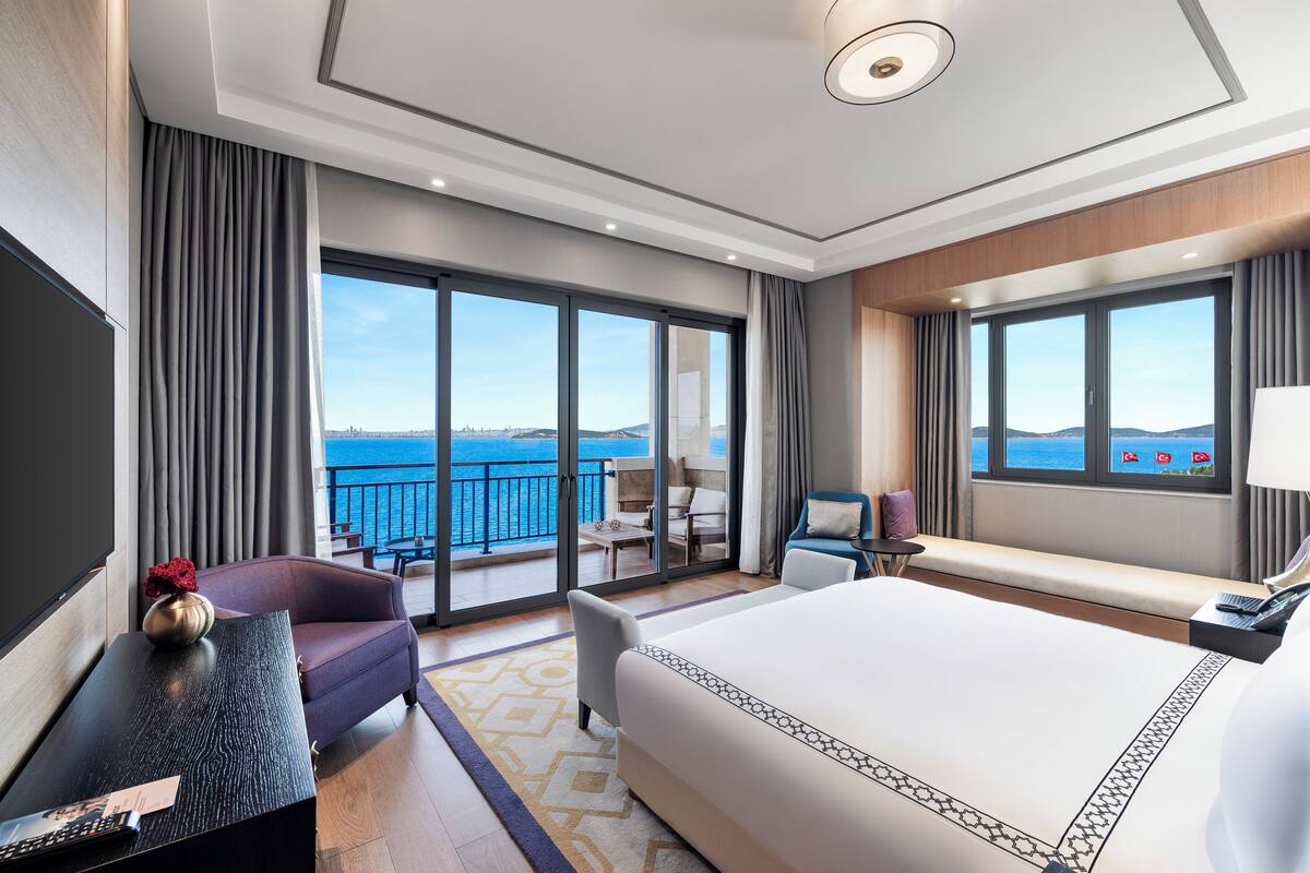 A modern hotel room with a large bed, a desk, a chair, and a balcony with a view of the sea.