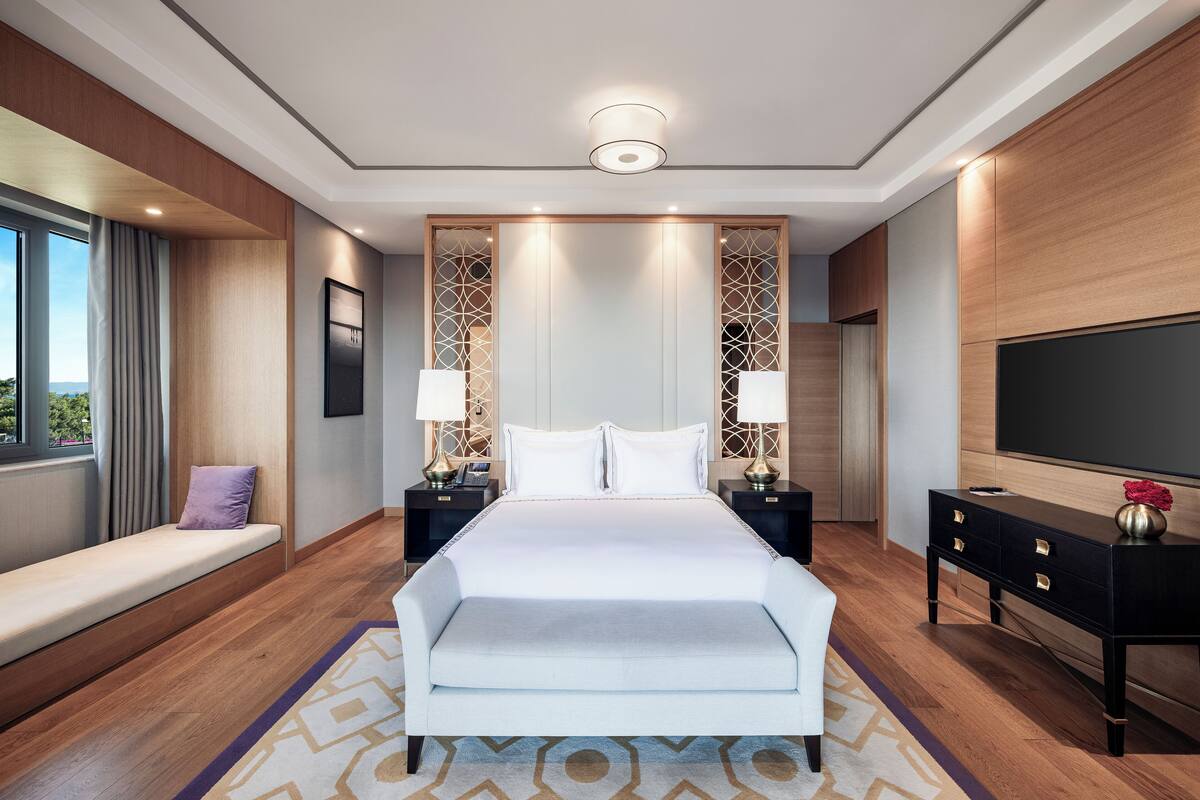 A modern hotel room with a large bed, bedside tables with lamps, a TV, and a view of the outdoors.