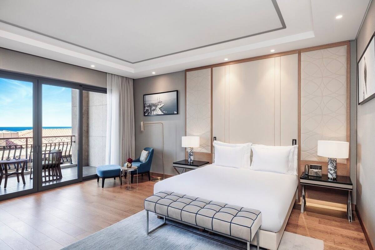 A modern hotel room with a large bed, a desk, a chair, a view of the beach, and a sliding glass door.
