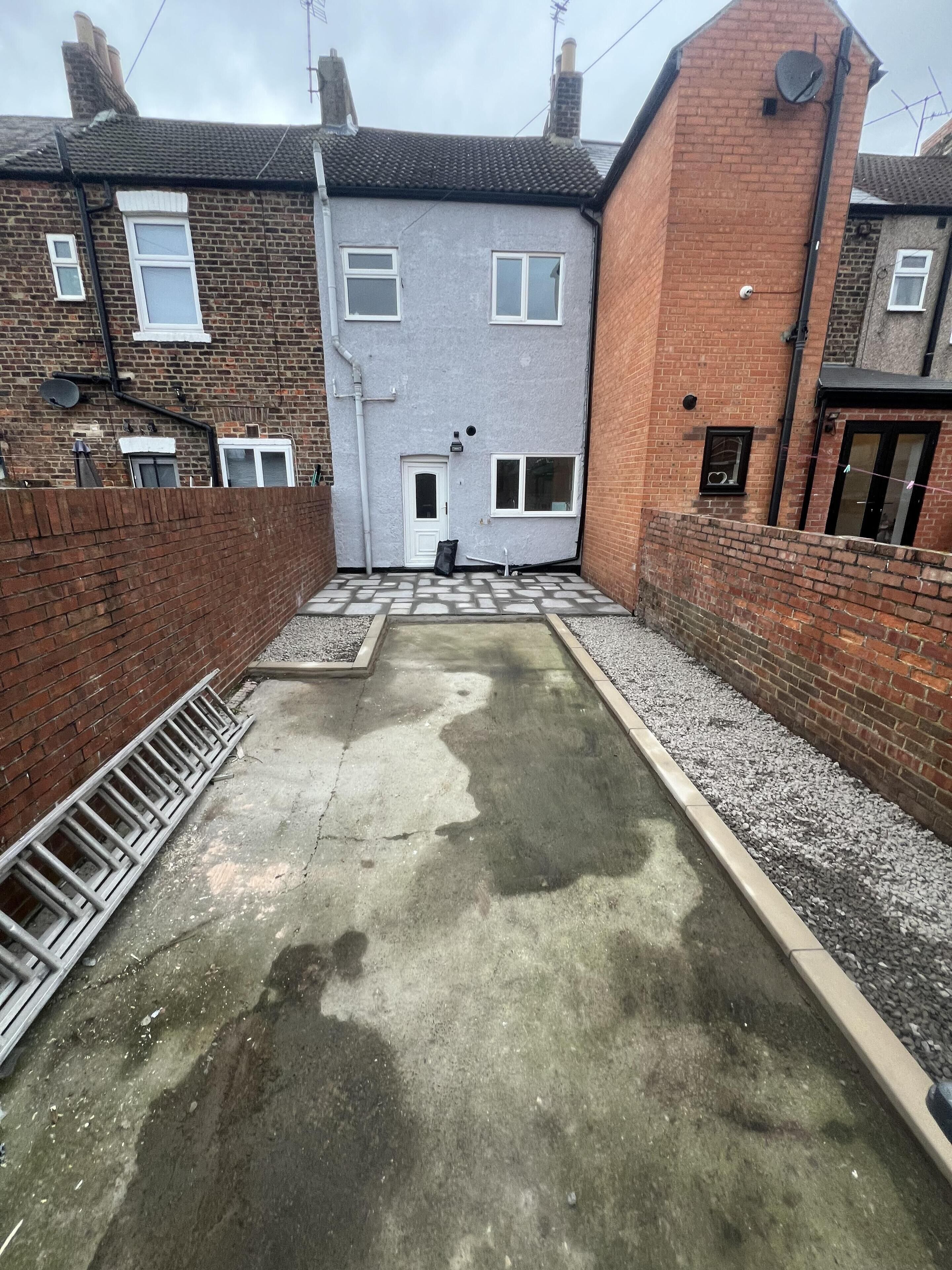 A narrow alleyway with a concrete path, brick walls, and a small garden area with gravel.