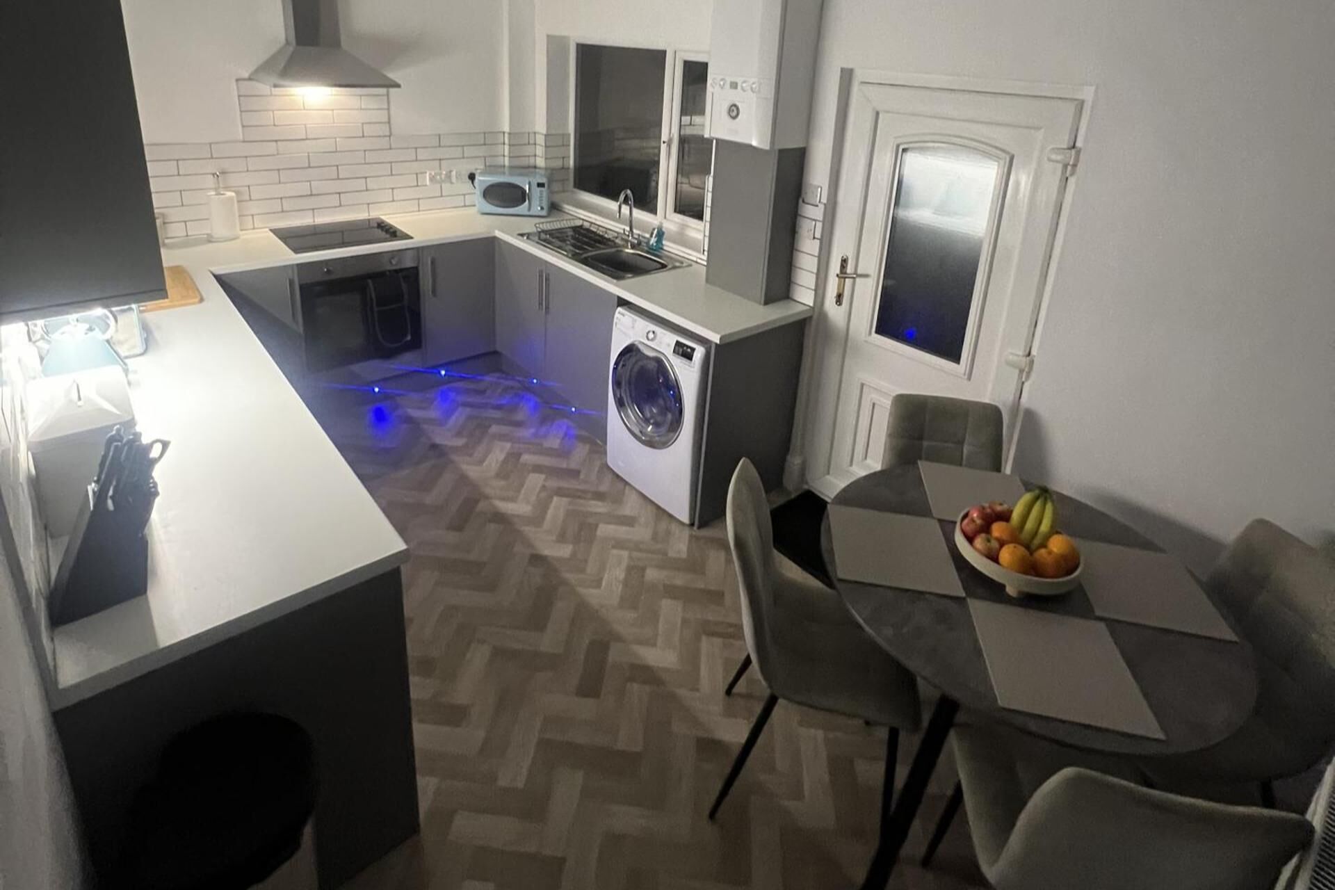 A compact kitchen with a washing machine, a microwave, and a dining area with a fruit bowl.