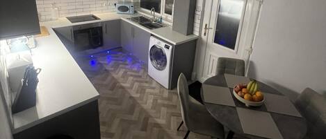 A compact kitchen with a washing machine, a microwave, and a dining area with a fruit bowl.