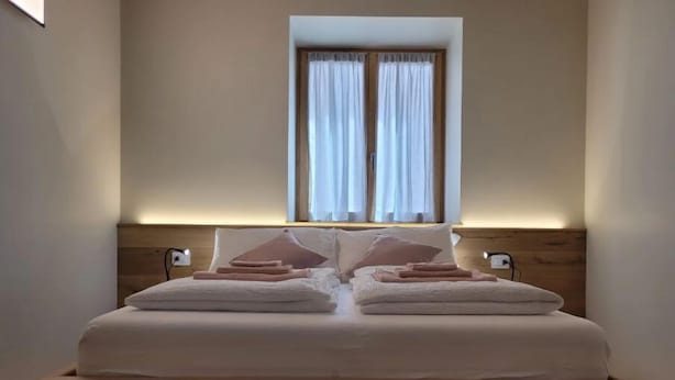 A neatly made bed with pillows and folded towels, a wooden headboard, and a window with curtains. - Magnolia LevicoGround floor apartment in the center of Levico Terme (Levico Terme)