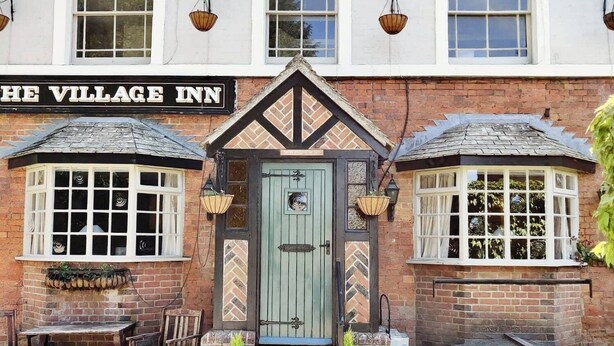 Perinteinen rakennus, jonka yläpuolella on kyltti, jossa lukee ”The Village Inn”. Siinä on vihreä ovi, kaksi erkkeri-ikkunaa ja pieni ulkona oleva oleskelualue, jossa on penkki ja kaksi ruukussa olevaa kasvia.