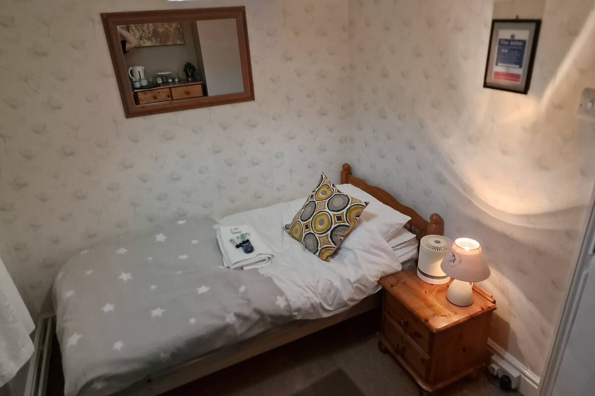 Un lit bien fait, recouvert d’une couvre-lit à motifs d’étoiles, une table de chevet en bois avec une lampe, et une petite commode en bois.