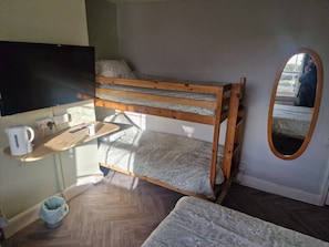 A bunk bed with a wooden frame and a mattress with a floral pattern. There is a small shelf with a kettle and a remote control. A small round chair is visible in the corner.
