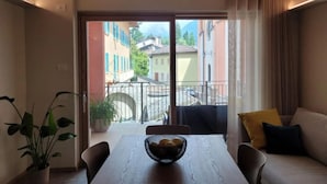 A dining area with a wooden table, chairs, and a bowl of bread. There is a balcony with a view of colorful buildings and a cobblestone street. - Magnolia LevicoApartment with terrace in the center of Levico Terme (Levico Terme)