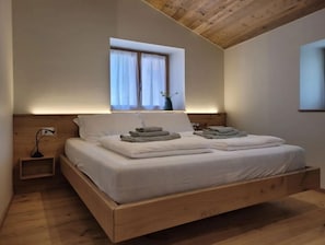 Une chambre à coucher avec un cadre de lit en bois, du linge de lit blanc, une table de chevet avec une lampe et une fenêtre avec des rideaux.