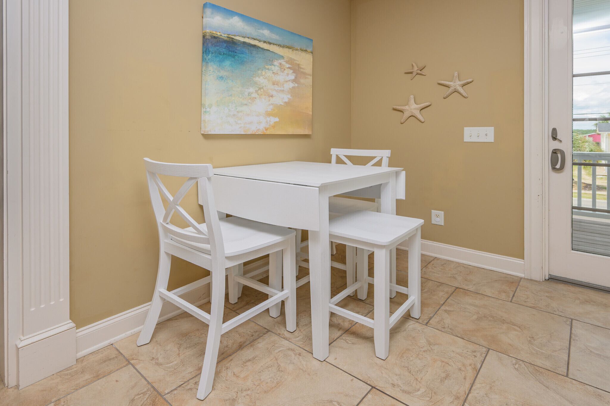 Un coin repas avec une table et des chaises blanches, un tableau sur le thème de la plage au mur et des décorations en forme d’étoiles de mer.