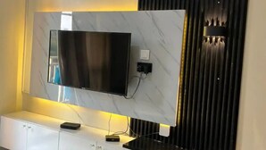 A modern living room with a large flat-screen TV mounted on a marble wall, a white cabinet below, and a black decorative panel to the right. - IvyRose Luxury Apartment 1 (Kampala)