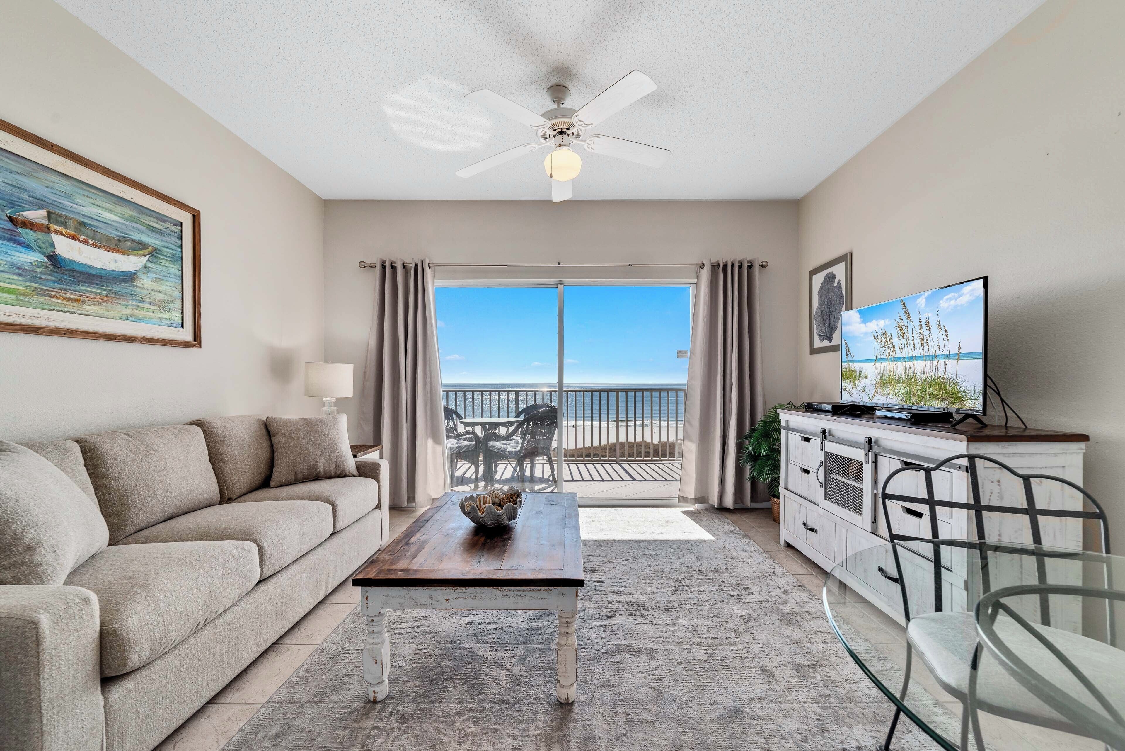Ein Wohnzimmer mit einer Couch, einem Couchtisch, einem Fernseher und einem Balkon mit Blick auf den Strand.