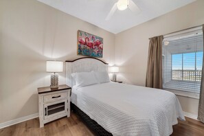 Un dormitorio con una cama grande, dos mesitas de noche con lámparas, un ventilador de techo, un cuadro de flamencos y una ventana con cortinas.
