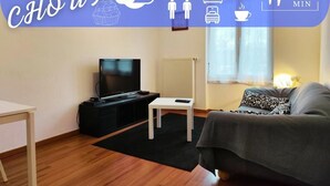 A modern living room with a flat-screen TV, a small white table, a sofa, and a window with natural light.