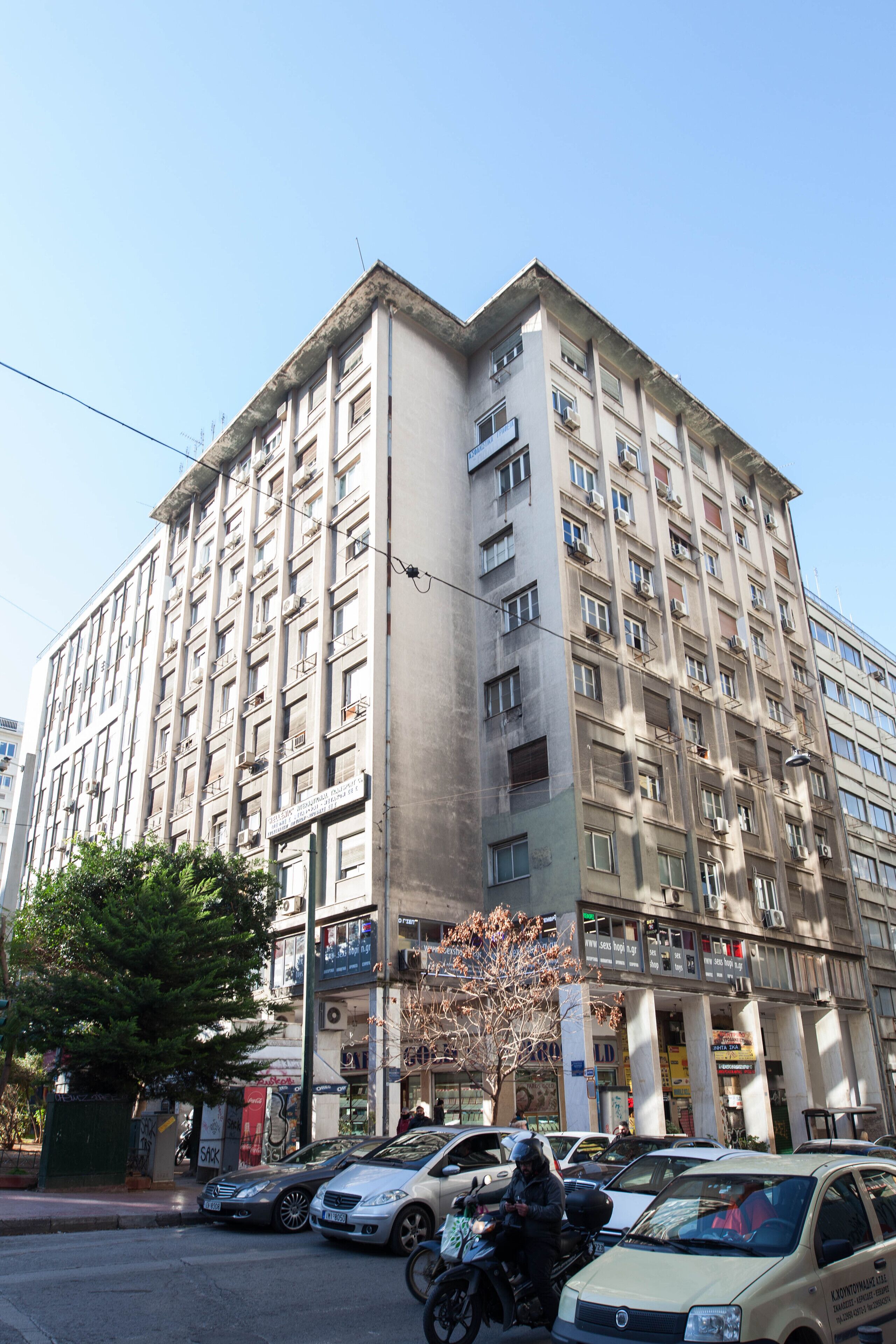 A multi-story building with a uniform facade, several balconies, and a parking area with cars and motorcycles.
