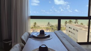 Un coin repas avec une table dressée pour deux personnes, donnant sur une plage bordée de palmiers et offrant une vue sur un ciel dégagé.