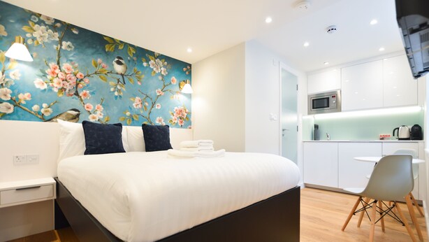 A modern hotel room with a large bed, a wall-mounted TV, a small kitchen area with a microwave, and a dining table with chairs. - Sale Place Serviced Apartments by Concept Apartments (London)