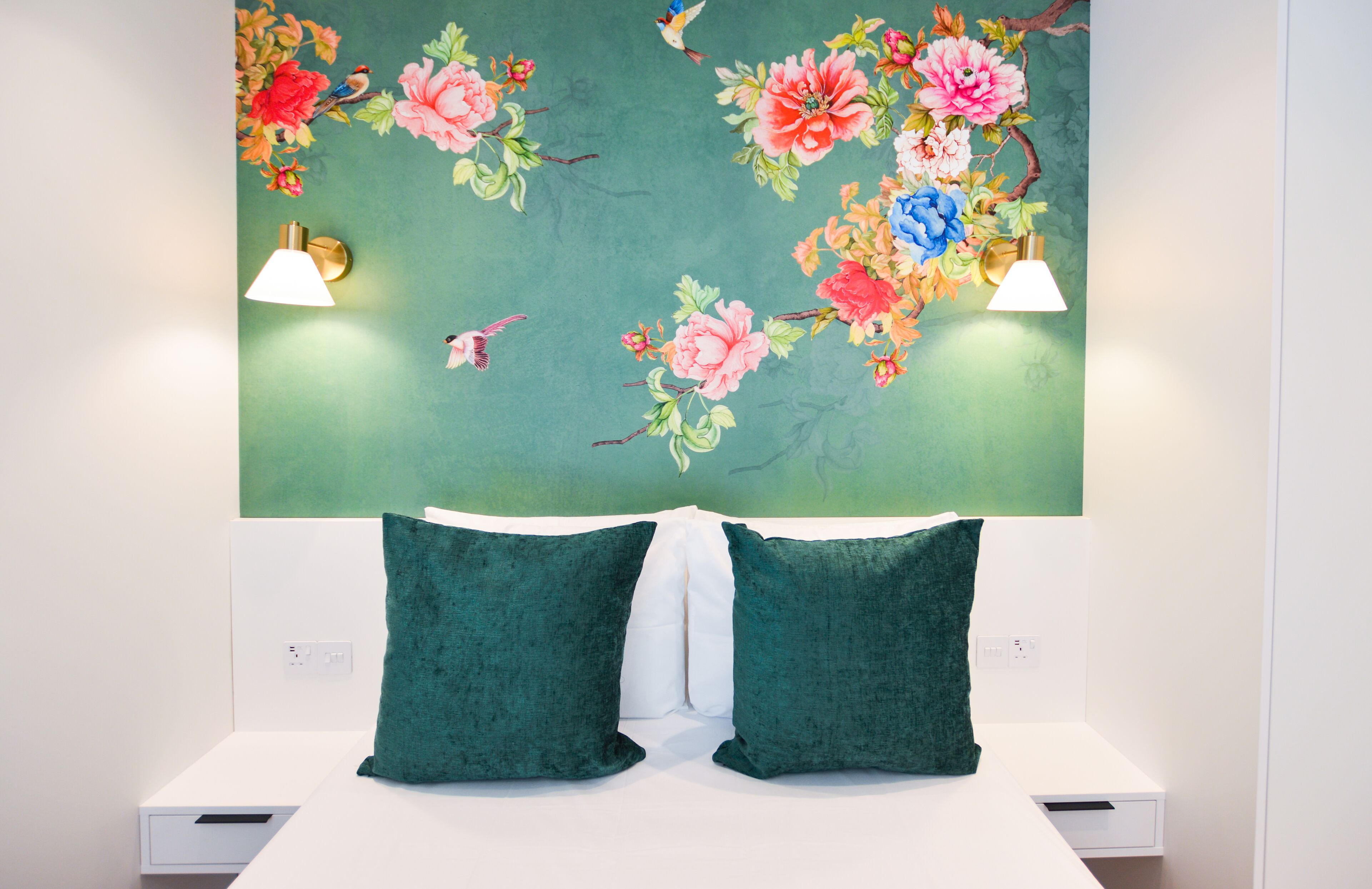 Une chambre à coucher avec un mur décoré d’un motif floral vert, deux coussins verts et des lampes de chevet.
