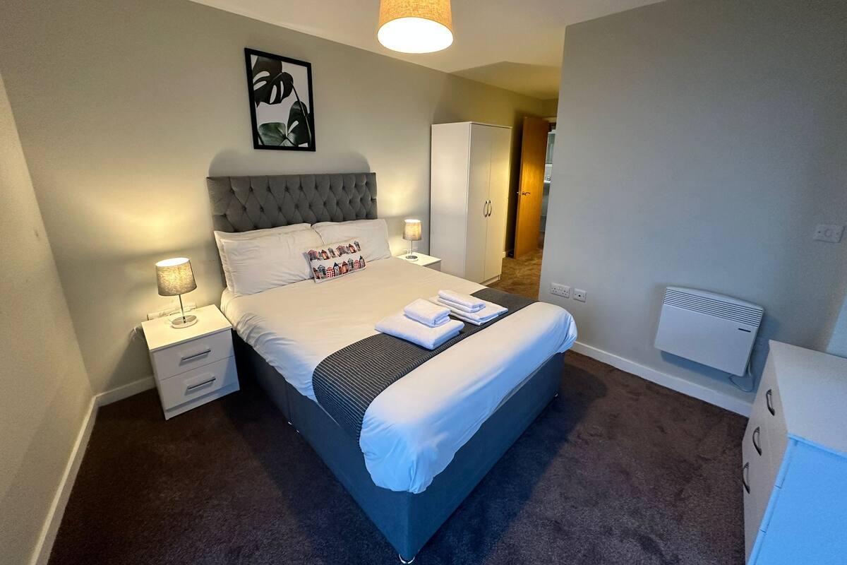 Une chambre à coucher bien aménagée, avec un grand lit, des lampes de chevet, une œuvre d’art accrochée au mur et une porte visible donnant sur une autre pièce.