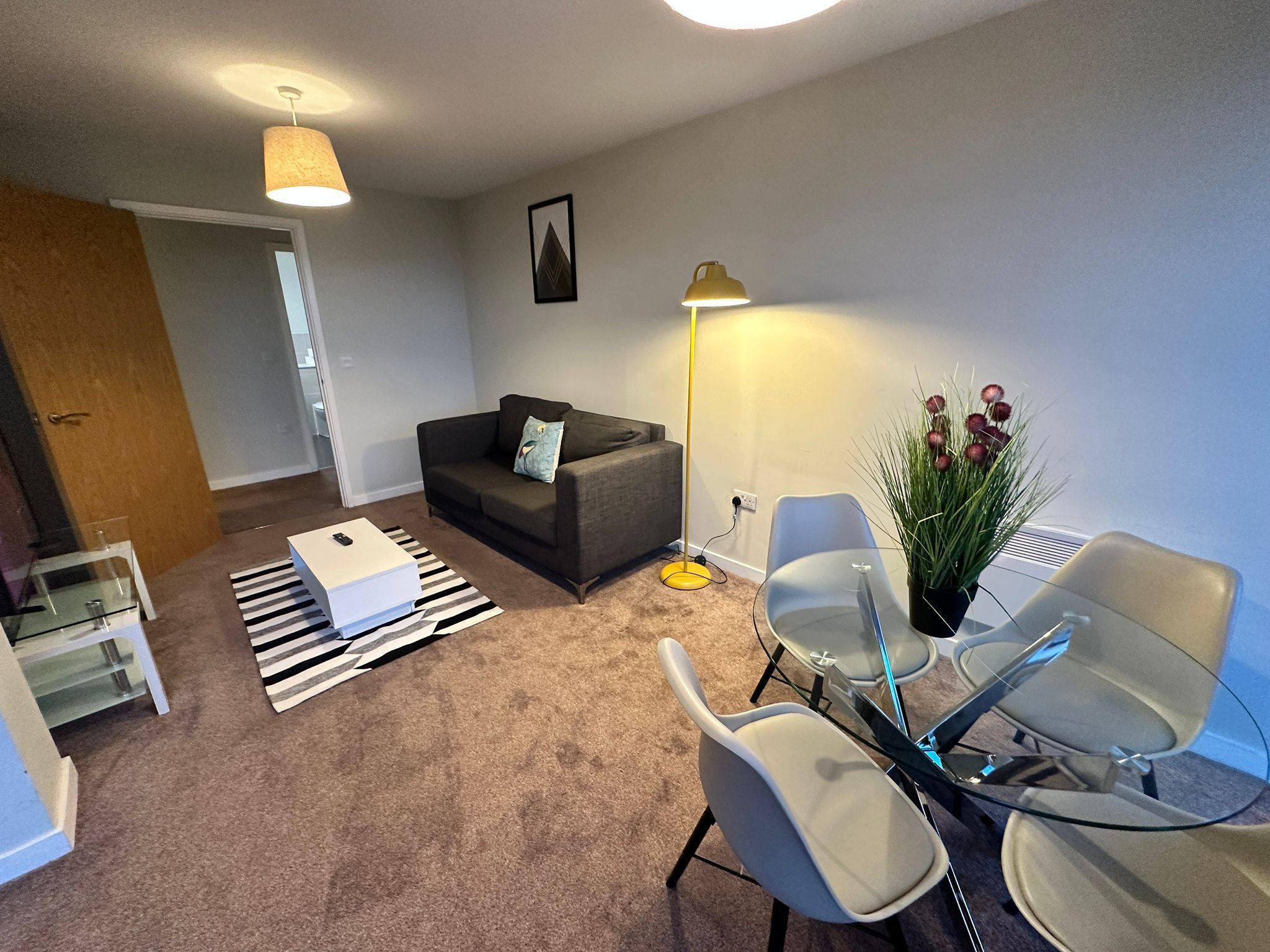 A modern living room with a sofa, a coffee table, a glass dining table with chairs, a floor lamp, and a potted plant.