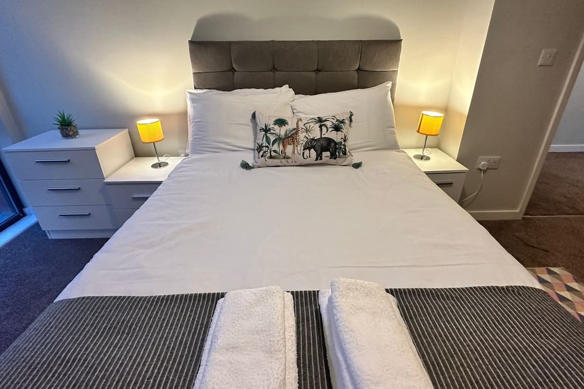 Un lit bien fait, doté d’une tête de lit capitonnée, de deux lampes de chevet, d’une petite plante et d’un coussin décoratif à motifs animaliers.