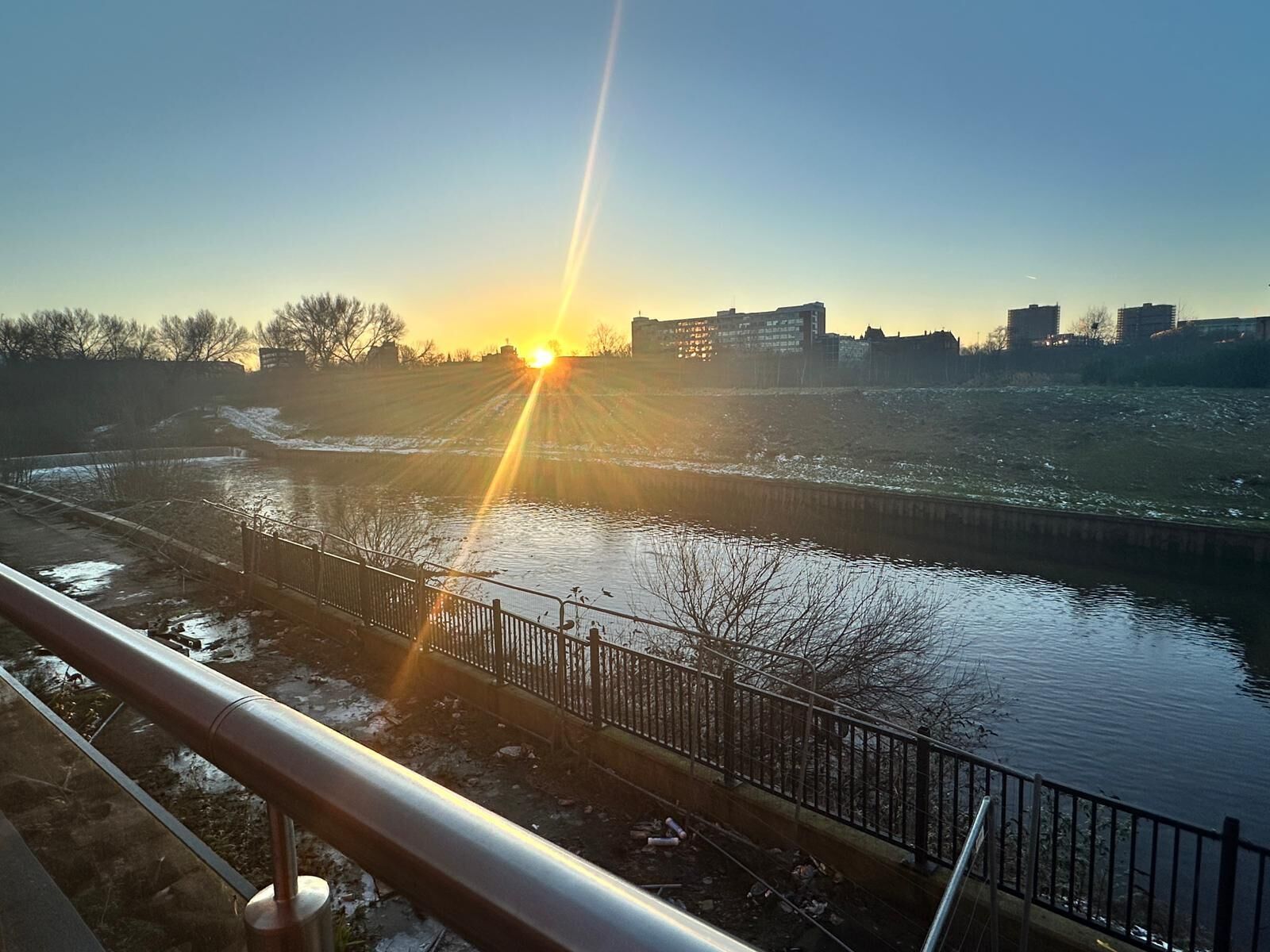 A river with a metal railing in the foreground, a clear sky, and buildings in the background.