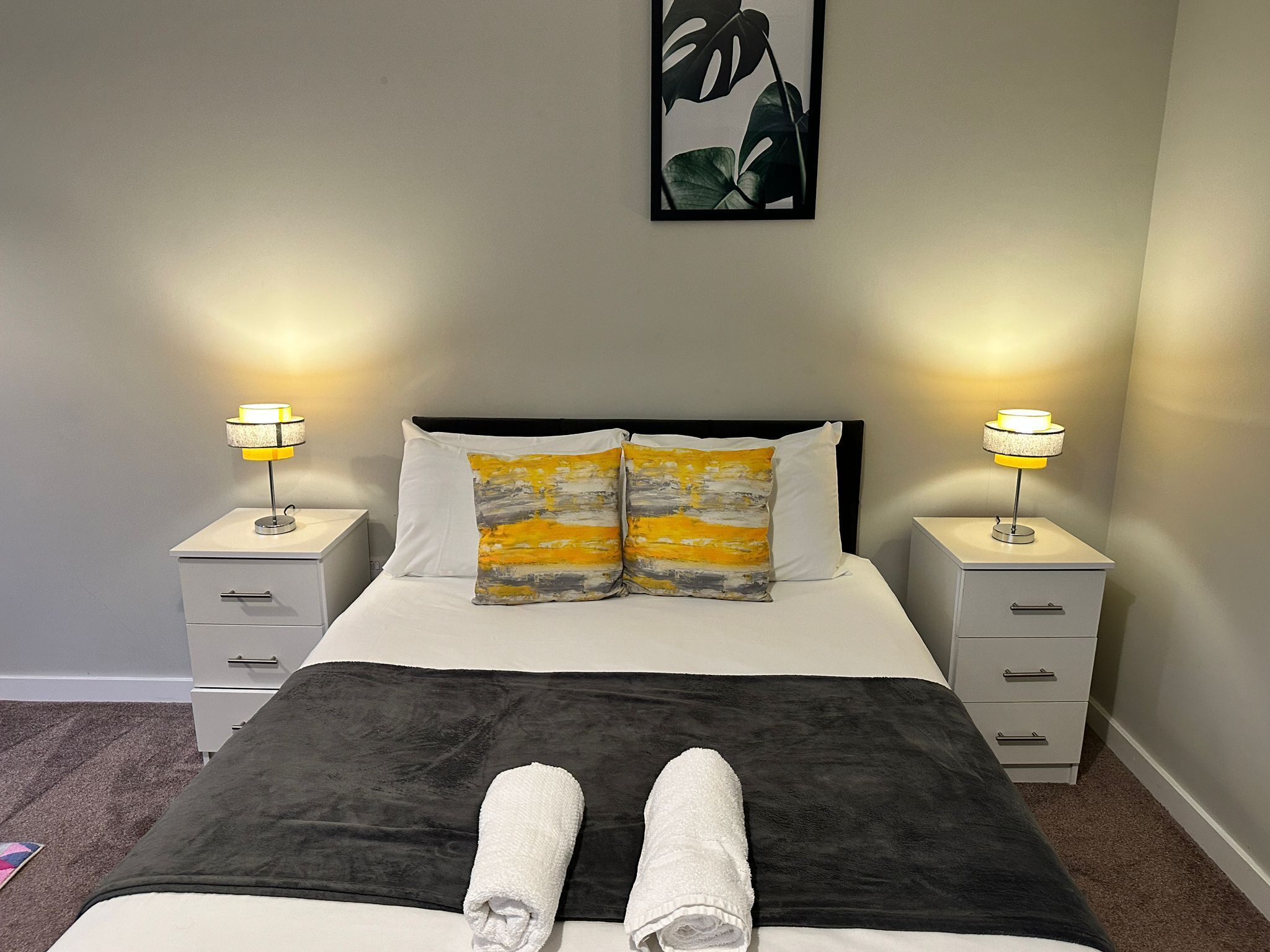 Ein ordentlich bezogenes Bett mit grau-weißer Decke, zwei Kissen mit gelb-grauem Muster und zwei Nachttischlampen.