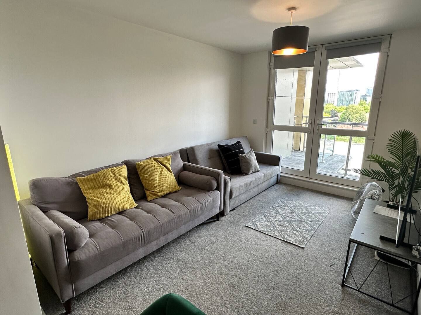 A modern living room with a grey sofa, yellow and black cushions, a grey carpet, a small table with a laptop, and a large window with a view of buildings and trees.